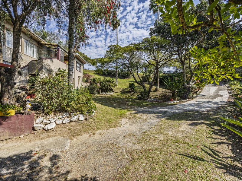 A view of the property's front exterior, featuring a concrete driveway, mature trees, and established garden landscaping leading toward the house entrance.