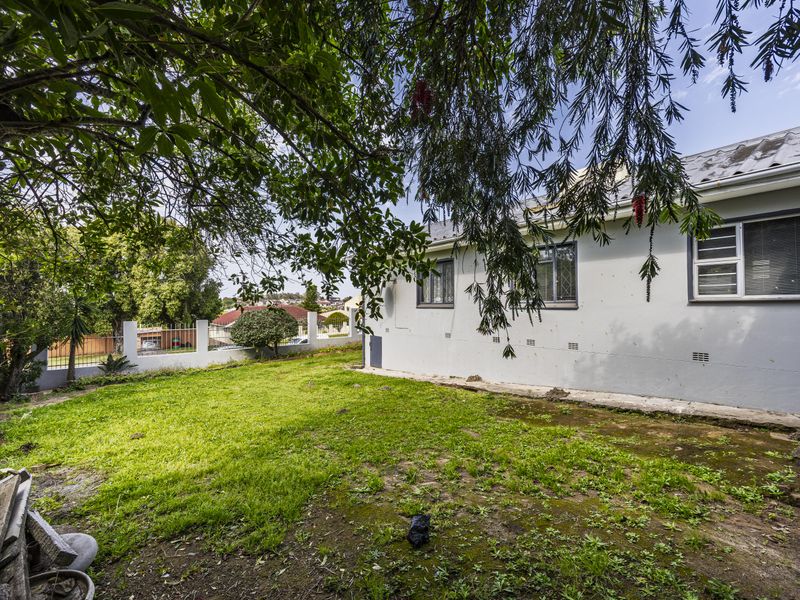 Rear exterior view of the house featuring a lawn area, boundary wall, and metal roofing with visible wear on the exterior walls and concrete.