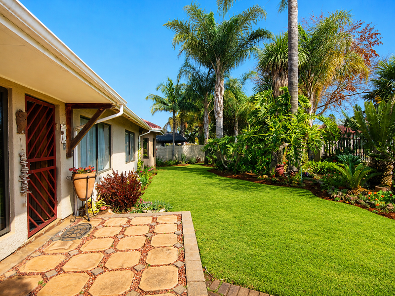 A well-maintained rear garden featuring a paved patio area, lush green lawn, and mature palm trees, providing a private outdoor living space.