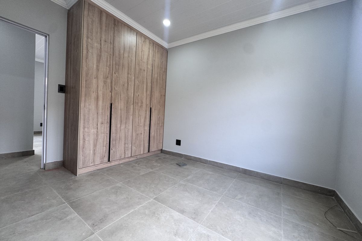 A clean, empty bedroom featuring modern wood-finish built-in cupboards, neutral tiled flooring, and crisp white walls with ceiling cornice detailing.