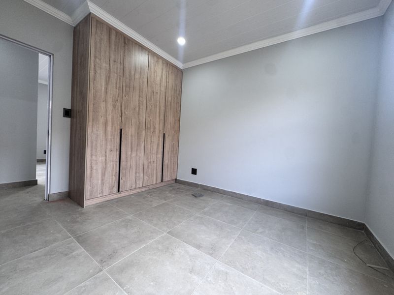 A clean, empty bedroom featuring modern wood-finish built-in cupboards, neutral tiled flooring, and crisp white walls with ceiling cornice detailing.