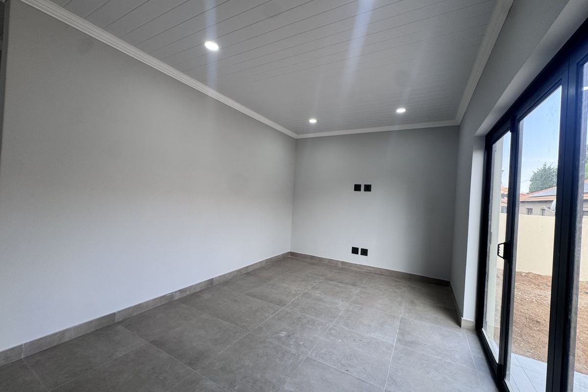 A bright, empty living room featuring modern grey floor tiles, recessed ceiling lighting, and large sliding glass doors providing access to the exterior.