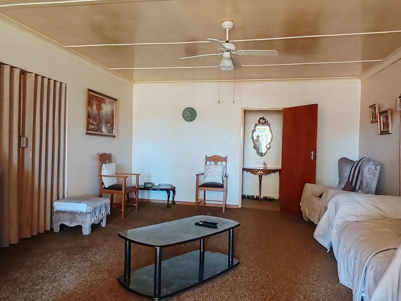 Spacious living room featuring a ceiling fan, panelled ceiling, and carpeted flooring, offering a dated interior ready for modern updates.
