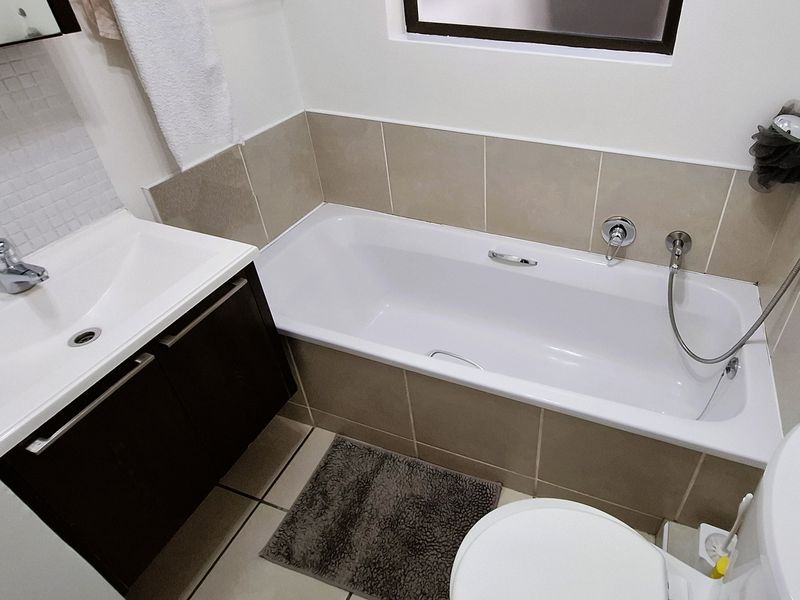 A compact, well-maintained bathroom featuring a modern vanity, bathtub with handheld shower attachment, and neutral wall and floor tiling.