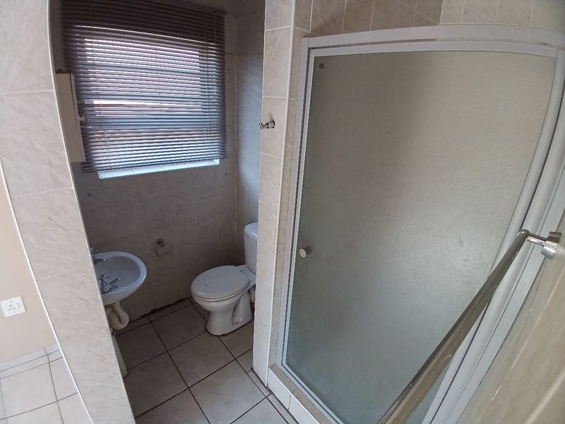 A functional bathroom featuring a shower enclosure, pedestal basin, and toilet with neutral tiled finishes and a window with a venetian blind.