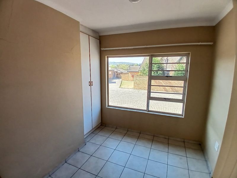 An empty bedroom featuring tiled flooring, a built-in wardrobe, and a large window with security bars, offering a view of the complex exterior.