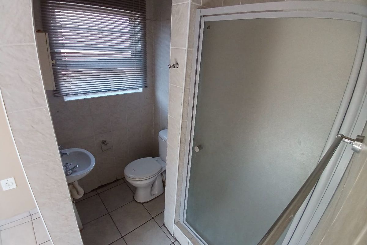A functional bathroom featuring a shower enclosure, pedestal basin, and toilet with neutral tiled finishes and a window with a venetian blind.