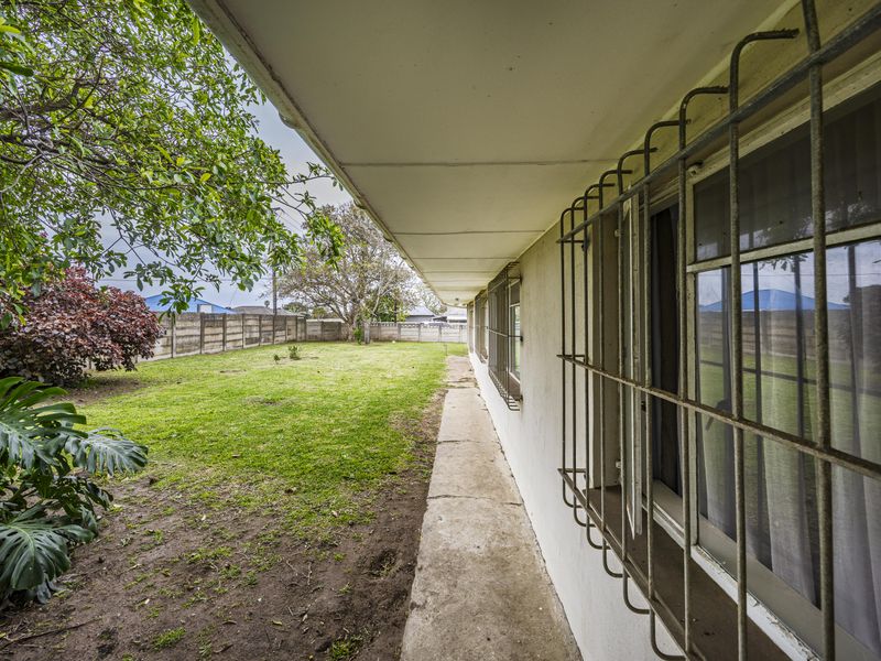 Rear exterior view of the property featuring a concrete walkway, security bars on windows, and a spacious garden enclosed by a precast boundary wall.
