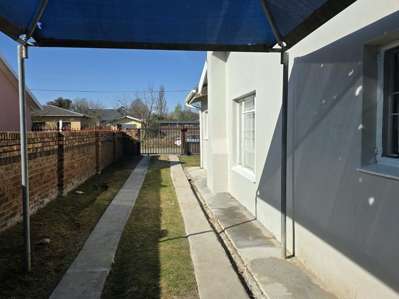 Side exterior view featuring a concrete driveway, brick boundary wall, and a covered carport area with security bars on the windows.