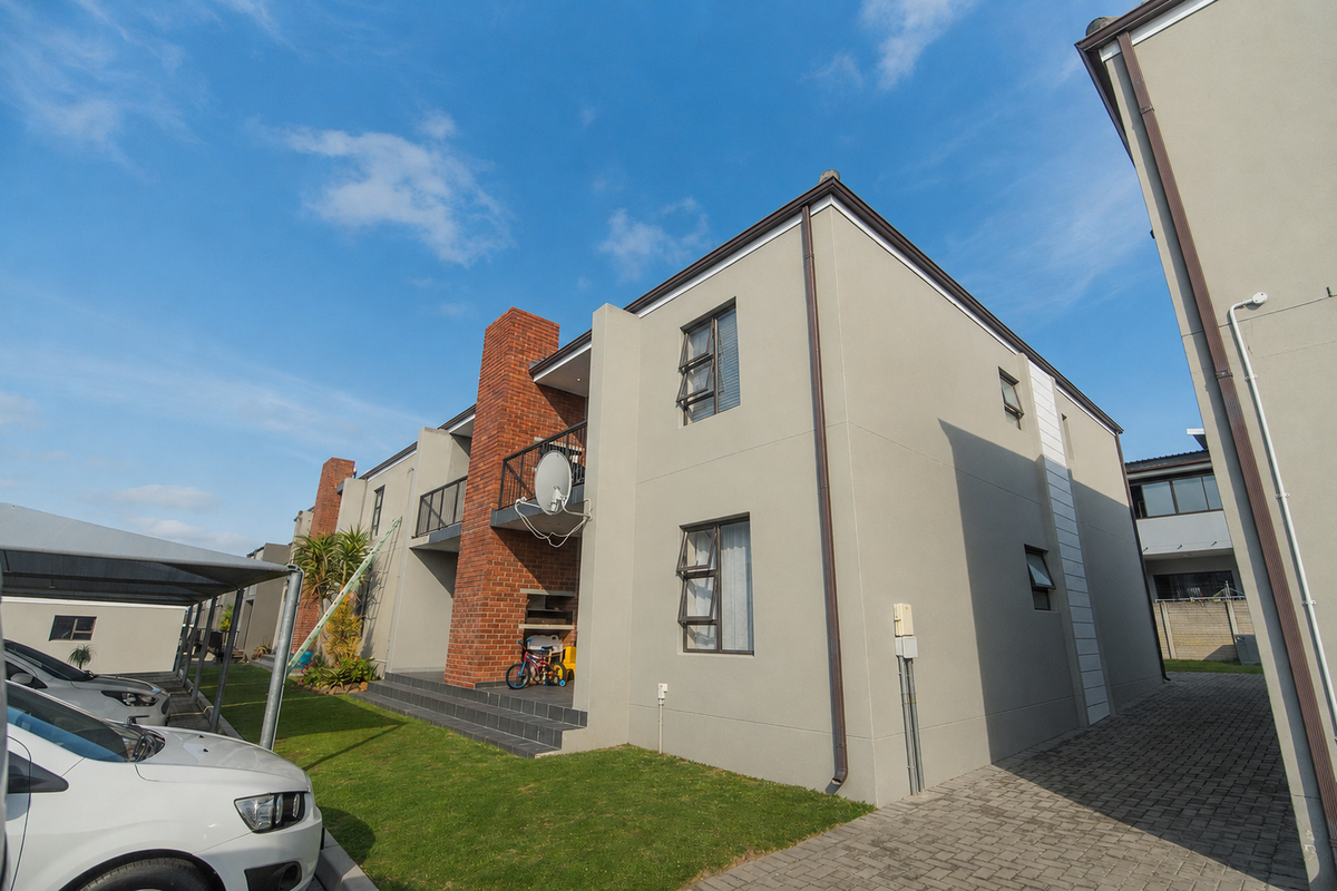 Rear view of a modern apartment complex featuring brick chimney detailing, a built-in braai area, balcony, and paved driveway access.