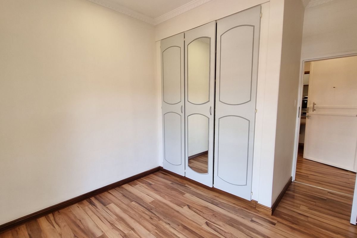 A clean, empty bedroom featuring light-toned laminate flooring, built-in cupboards with a central mirror panel, and decorative ceiling cornicing.
