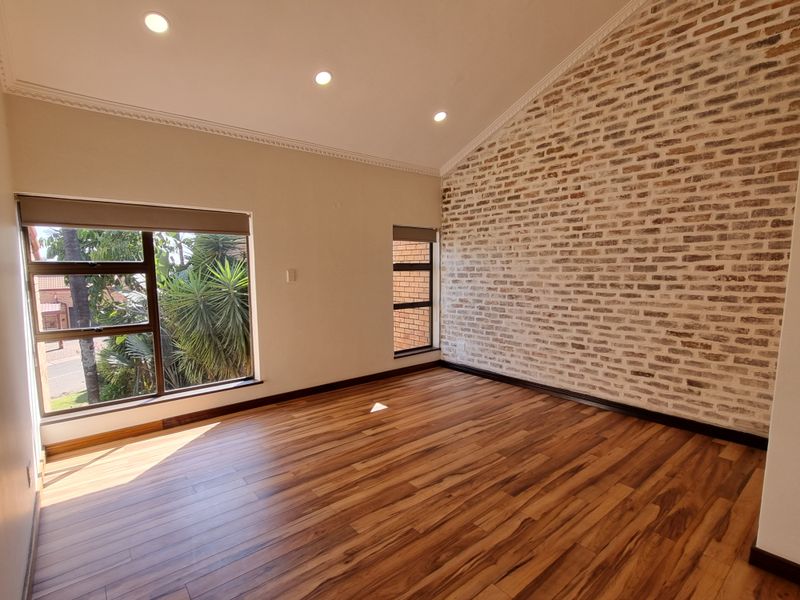 Bright and spacious living area featuring a striking exposed brick feature wall, modern laminate flooring, and large windows with roller blinds.