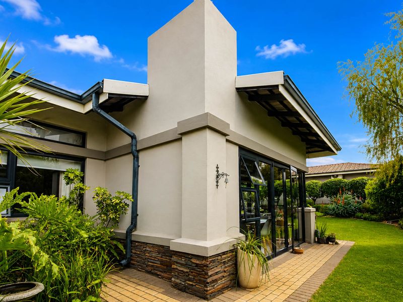 A well-maintained rear exterior view featuring stone cladding, modern aluminium-framed windows, a chimney stack, and a paved patio area overlooking a landscaped garden.