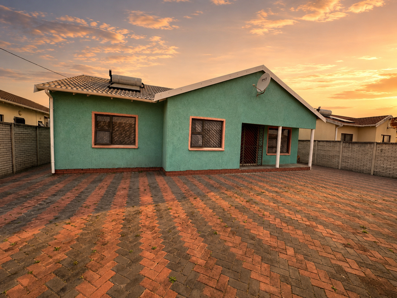 A front exterior view of a single-story house featuring a paved driveway, solar water heating system, and perimeter walling at sunset.
