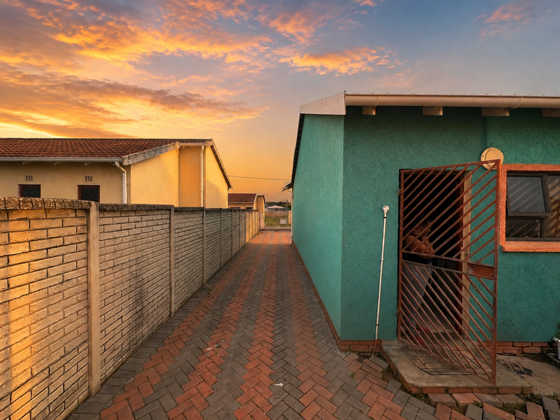 A view of the side passage between two residential properties, featuring paved ground, a brick boundary wall, and a teal-colored exterior wall with security gate.