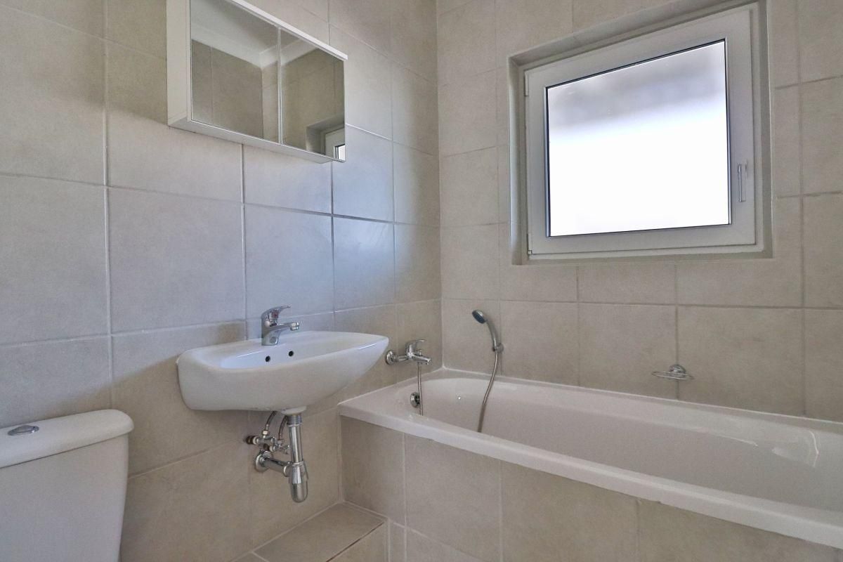 A clean, functional bathroom featuring a wall-mounted basin, bathtub with handheld shower, and a mirrored medicine cabinet with neutral wall tiling.
