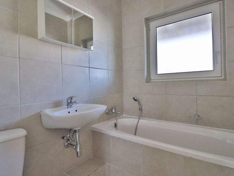 A clean, functional bathroom featuring a wall-mounted basin, bathtub with handheld shower, and a mirrored medicine cabinet with neutral wall tiling.