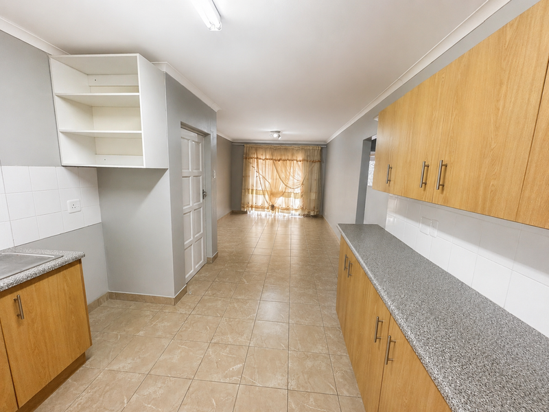 An open-plan kitchen and living area featuring light wood-look cabinetry, granite-effect countertops, and neutral ceramic floor tiles in a clean, vacant space.