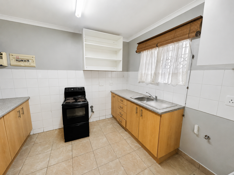 A functional kitchen featuring light wood cabinetry, laminate countertops, a stainless steel sink, and a freestanding electric stove with tiled splashbacks and flooring.