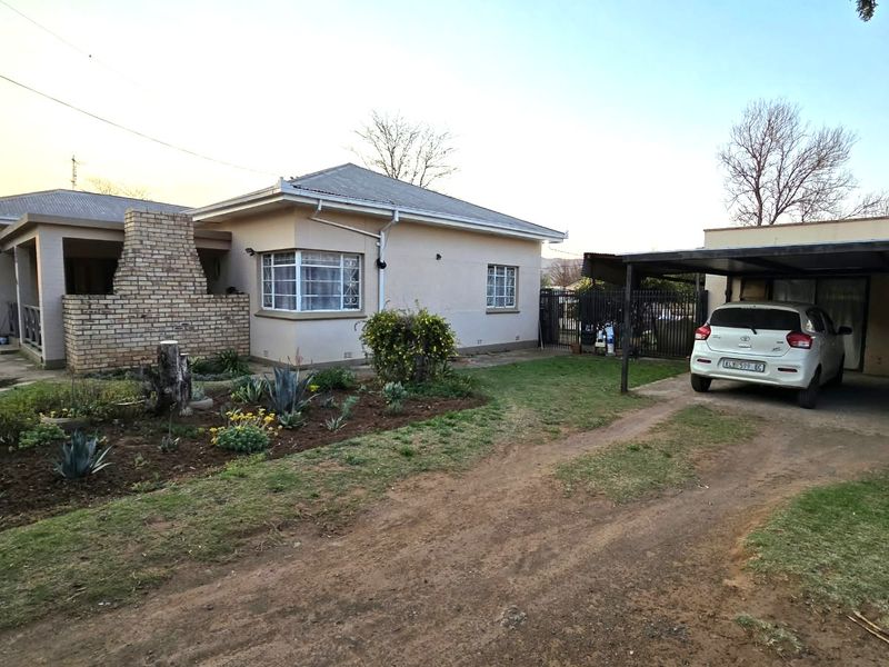 Front view of a single-story house featuring a brick chimney, metal roof, and an attached carport with a gravel driveway.