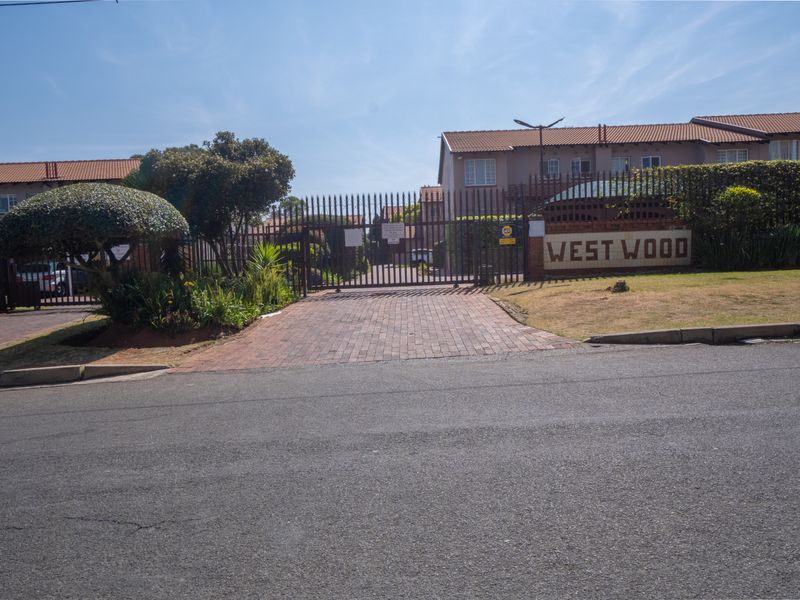 Street view of the West Wood townhouse complex entrance, featuring a secure gated access point, brick driveway, and established perimeter fencing.