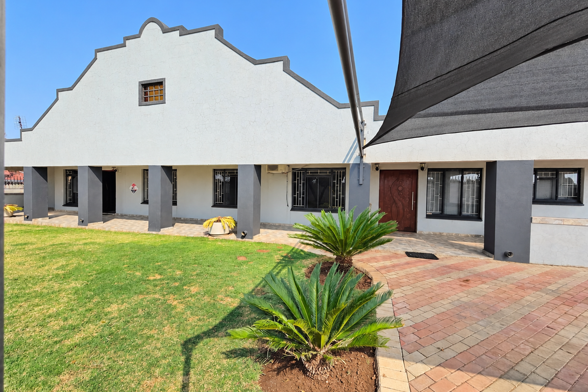 Rear view of the property featuring a gabled facade, paved patio area, shade sail, and well-maintained lawn with established landscaping.