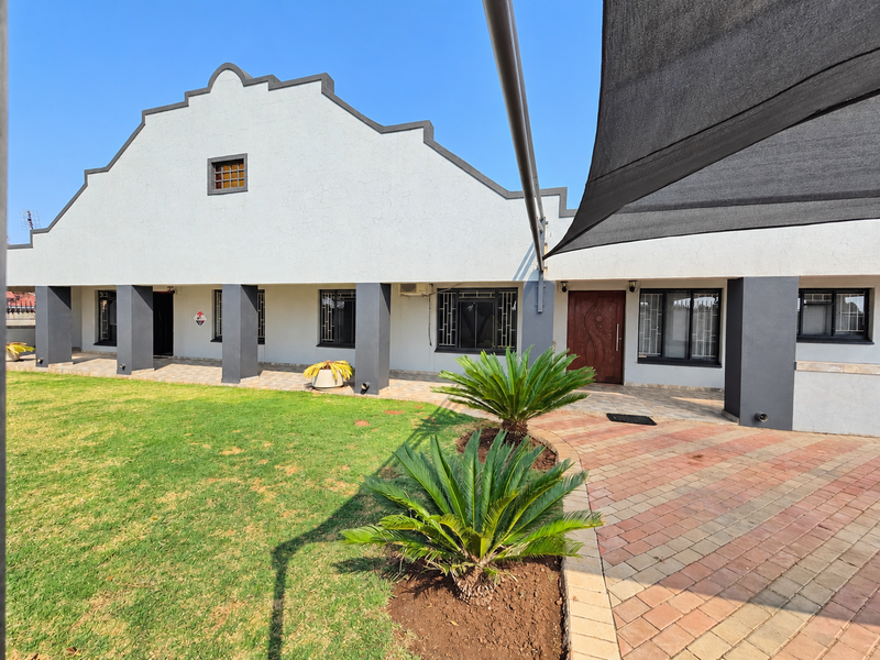 Rear view of the property featuring a gabled facade, paved patio area, shade sail, and well-maintained lawn with established landscaping.