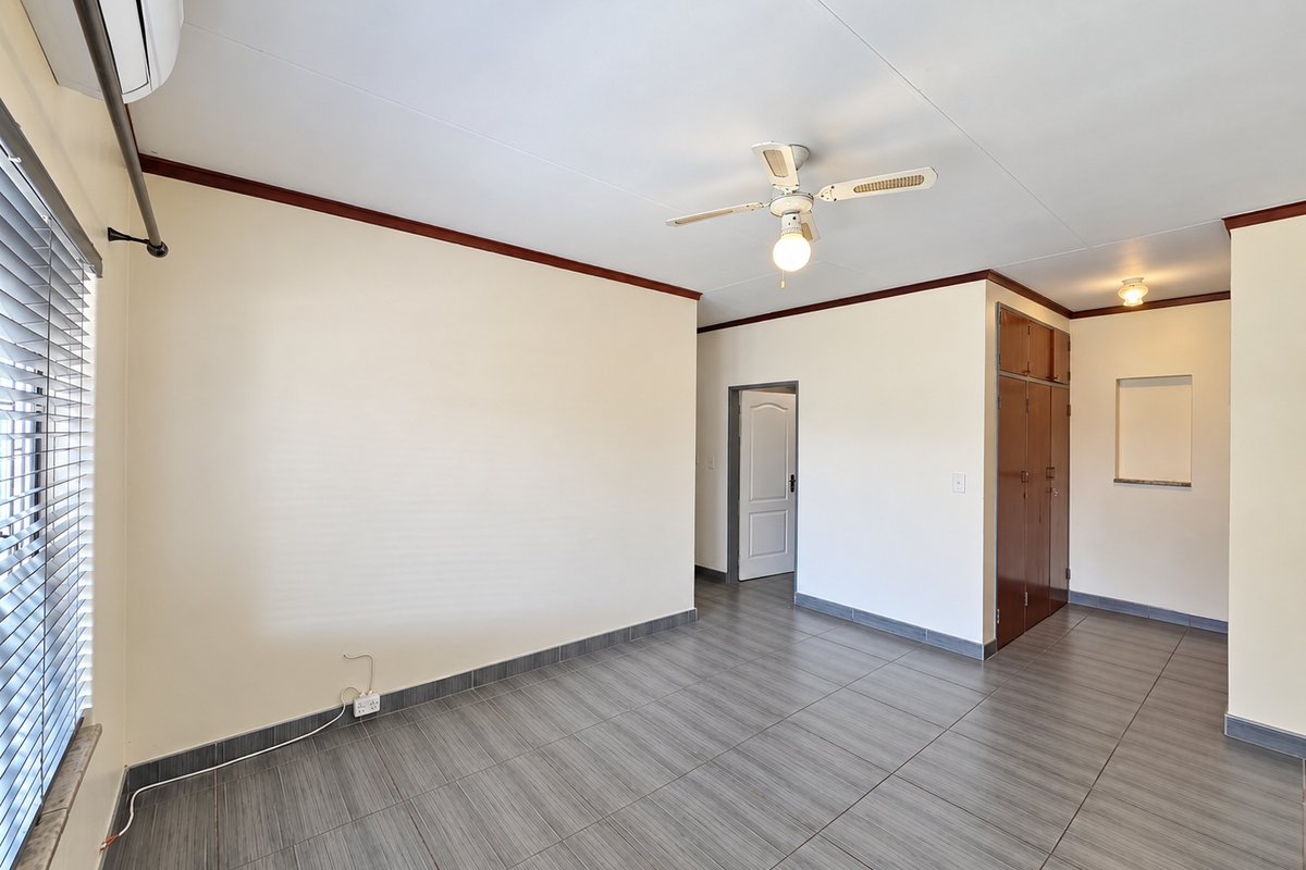 A spacious, empty bedroom featuring tiled flooring, built-in cupboards, a ceiling fan, and an air conditioning unit.