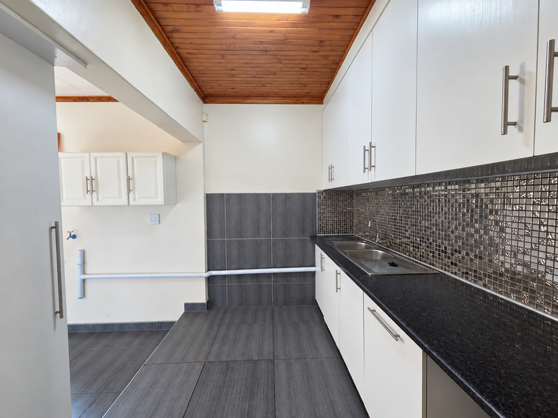 A clean and functional scullery featuring modern white cabinetry, dark granite countertops, a mosaic tile backsplash, and a warm timber ceiling.