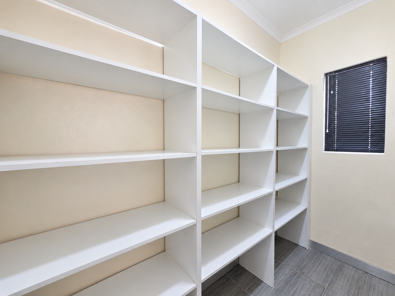A clean utility space or storage room featuring extensive built-in white shelving, tiled flooring, and a window with fitted venetian blinds.