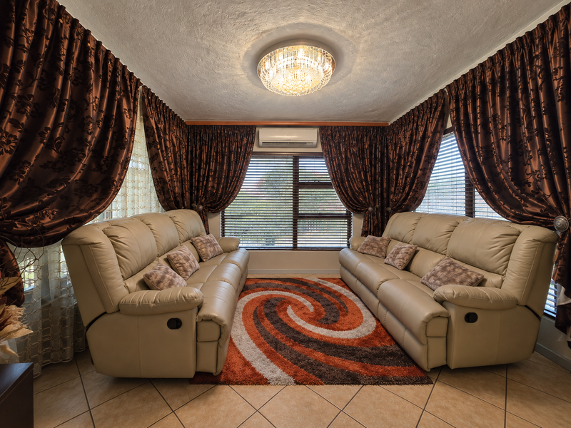 A well-maintained living room featuring tiled flooring, a central crystal chandelier, and a wall-mounted split-system air conditioner with large windows and venetian blinds.