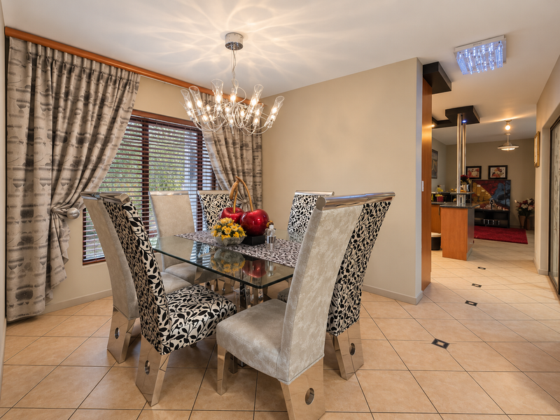 A well-lit dining area featuring ceramic floor tiles, modern chandelier lighting, and large windows with venetian blinds, leading into an open-plan kitchen space.