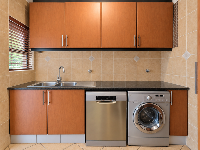 A functional laundry area featuring built-in wooden cabinetry, a stainless steel sink, tiled backsplash, and dedicated spaces for laundry appliances.
