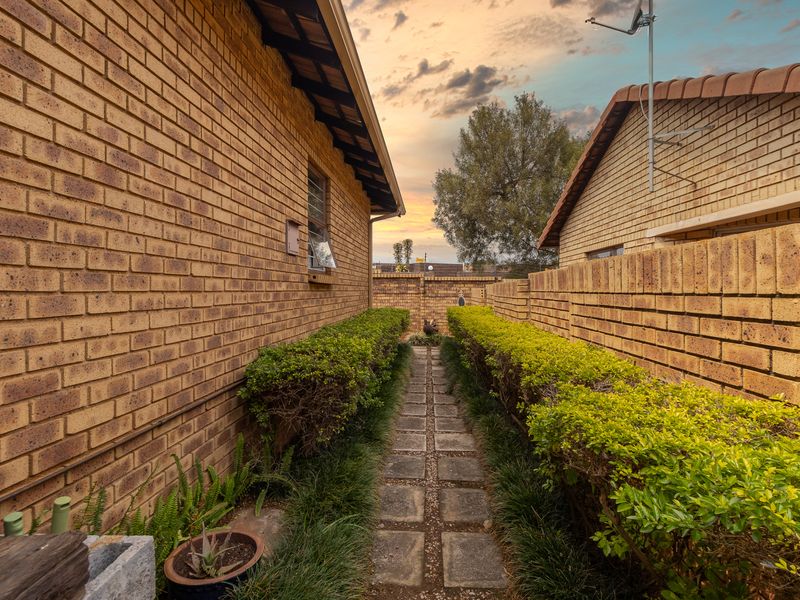 A narrow side passage between two brick townhouse structures featuring a paved walkway and manicured hedges.
