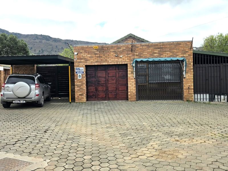 Front exterior view of a townhouse featuring a brick facade, a single garage, a covered carport, and a paved driveway with security fencing.