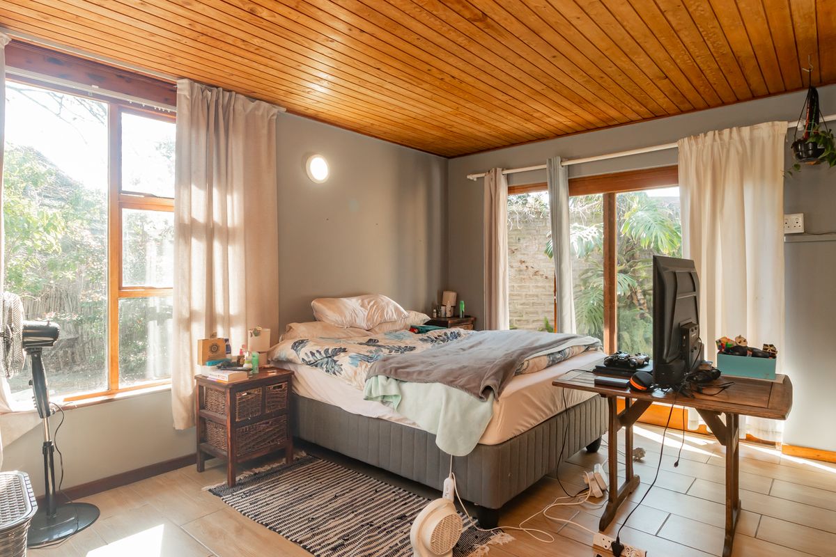 A bedroom featuring a warm timber-clad ceiling, tiled flooring, and large windows providing natural light and access to the garden area.