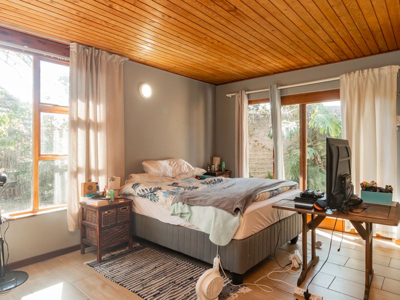 A bedroom featuring a warm timber-clad ceiling, tiled flooring, and large windows providing natural light and access to the garden area.