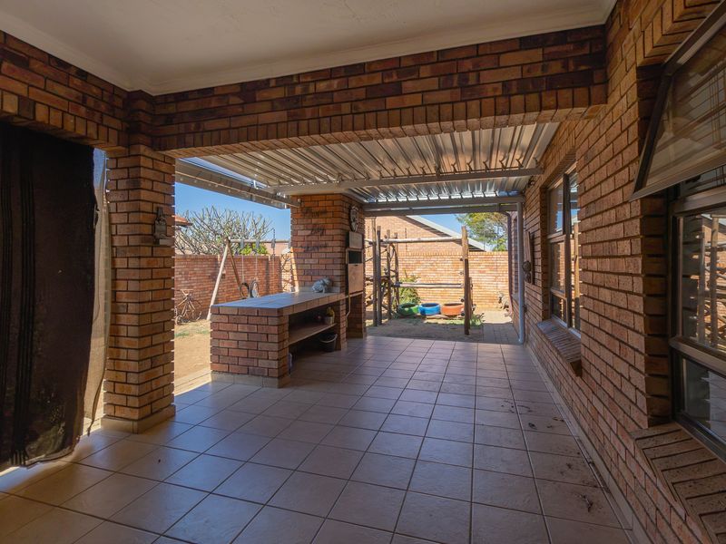 A covered outdoor patio featuring brick construction, tiled flooring, and a built-in braai area, overlooking the rear garden space.