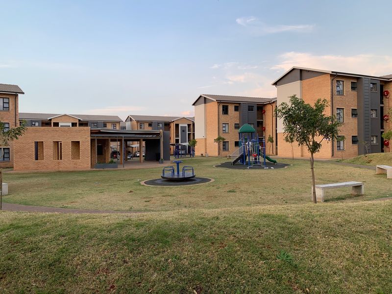 A well-maintained communal garden area within an apartment complex, featuring brick buildings, paved pathways, and a children's playground.