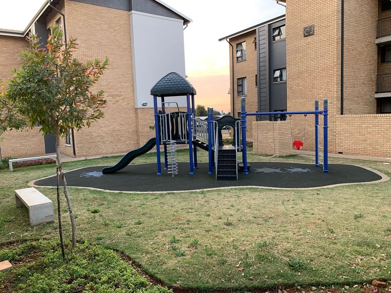 A well-maintained communal playground area situated between apartment blocks, featuring modern play equipment, rubberized flooring, and concrete seating within a landscaped garden setting.