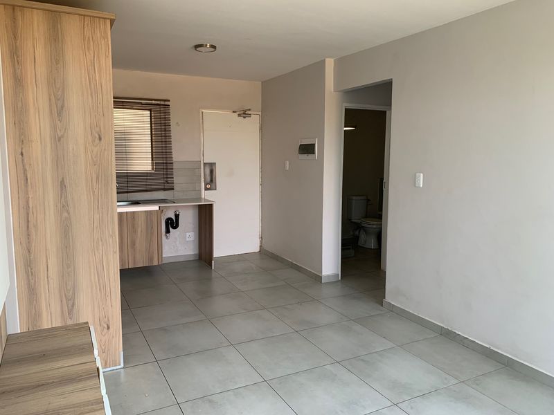 Open plan living area featuring tiled flooring, a basic kitchenette with exposed plumbing, and direct access to an adjacent bathroom.