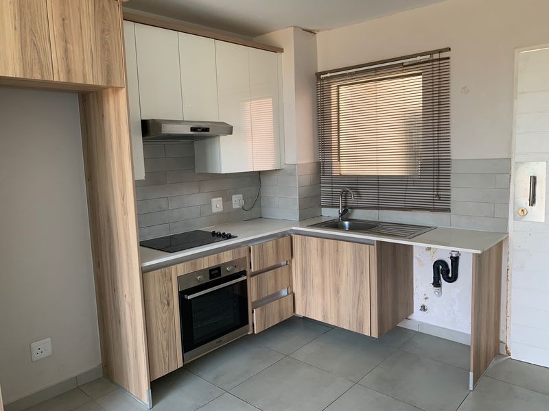 A compact kitchen featuring wood-look cabinetry, a built-in oven and hob, and a tiled backsplash, currently requiring minor repairs to the under-sink area.