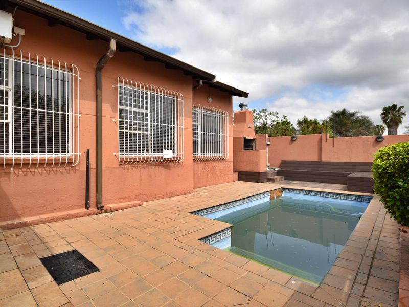 Rear exterior area featuring a swimming pool, paved patio, and a built-in braai, enclosed by a boundary wall with security bars on windows.
