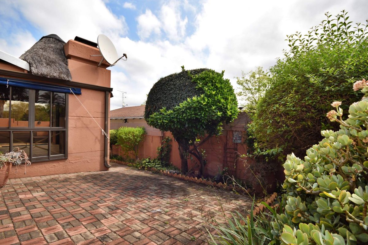 A rear exterior view featuring a paved patio area, a thatched roof section, and established garden landscaping against a boundary wall.