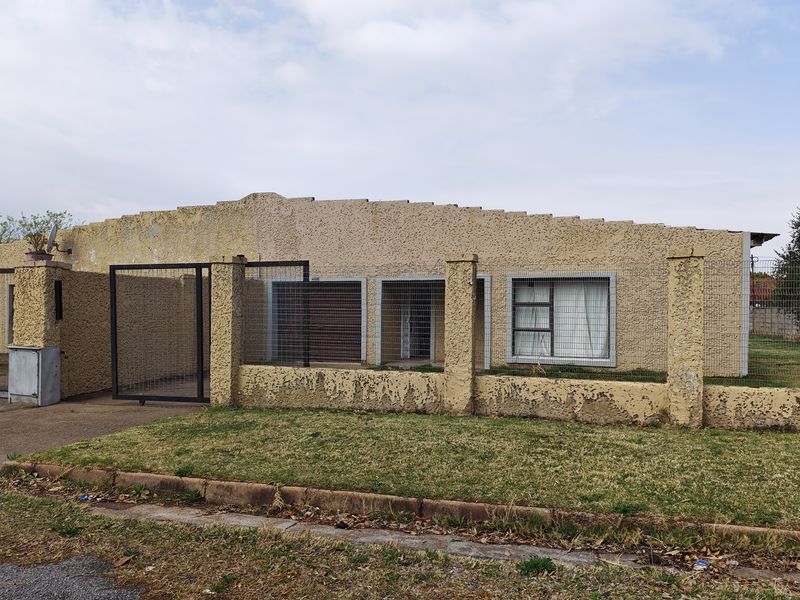 Front elevation of a single-story house featuring a textured stucco facade, perimeter security fencing, and a sliding gate entrance.