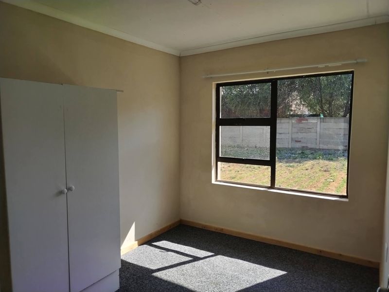 A simple, empty bedroom featuring carpeted flooring, neutral walls, and a large aluminium-framed window overlooking the rear garden.