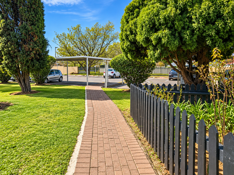 A well-maintained front garden area featuring a paved walkway, a picket fence, and a covered carport for vehicle parking.