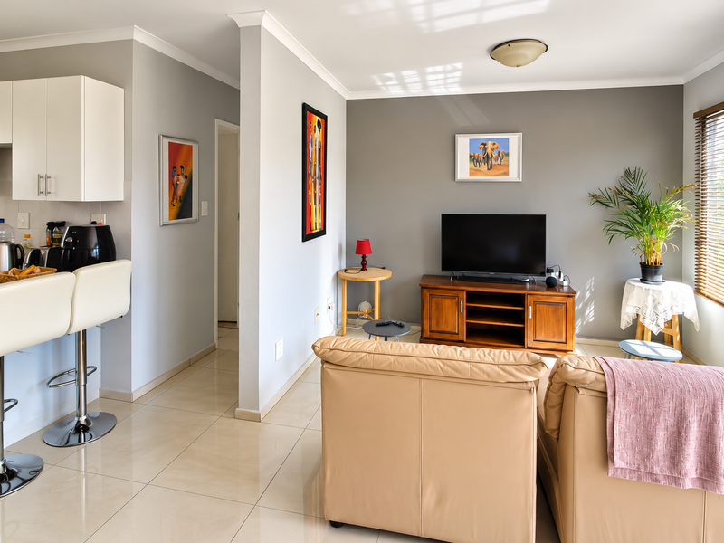 An open-plan living area featuring tiled flooring, a breakfast bar with kitchen cabinetry, and natural light through venetian blinds.