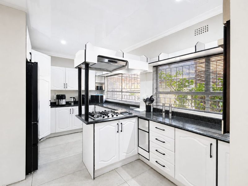 A well-maintained kitchen featuring white cabinetry, dark granite countertops, a gas hob with an extractor fan, and large windows with venetian blinds.
