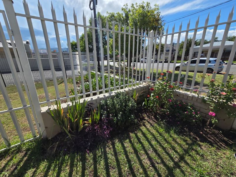 A view of the property's front boundary featuring a white metal security fence, a low brick retaining wall, and a landscaped garden bed.
